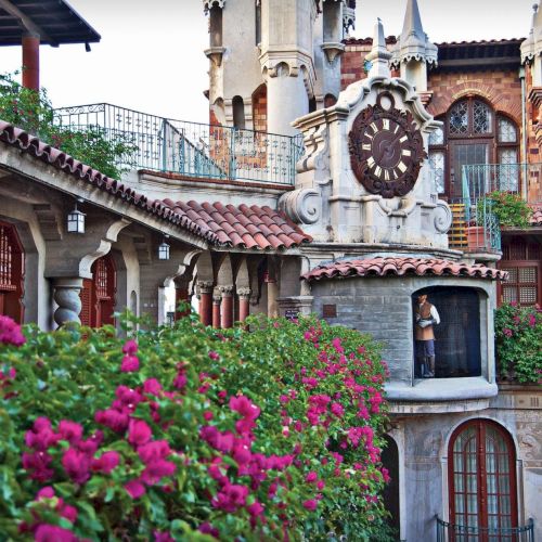A landscaped courtyard of ornate European-style buildings with an arched clock, pink flowers, and a man by a doorway.
