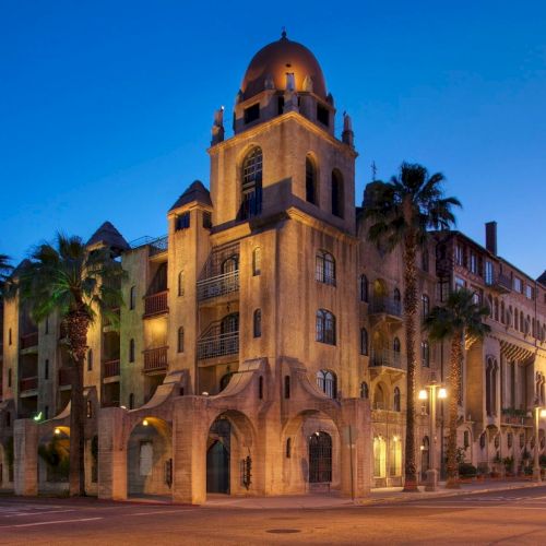 An illuminated historic church-like building with a large dome and arched walls, framed by palm trees at dusk.