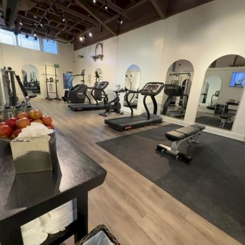 A bright gym with treadmills and cardio machines, mirrors, and a bench piled with oranges and apples.