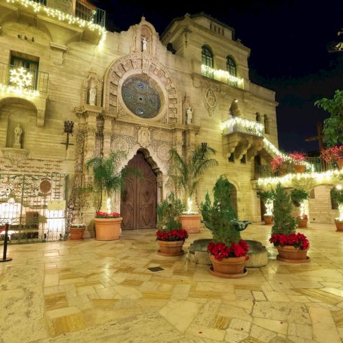 A warmly lit stone cathedral courtyard at night, decorated with string lights, potted plants, and ornate doors in front.