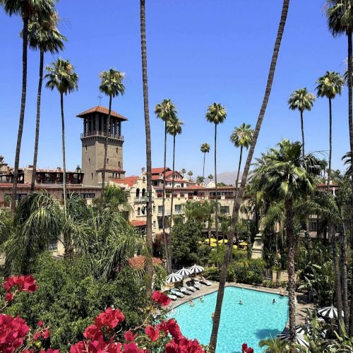A sunny tropical resort courtyard with palm trees, bright gardens, and a turquoise swimming pool surrounded by buildings.