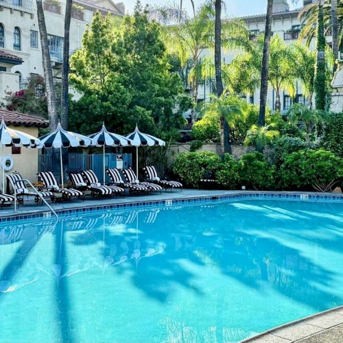 A resort pool with striped loungers, shade umbrellas, tall palms, and white buildings in a lush courtyard.