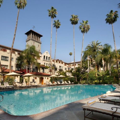 Sunny hotel courtyard with a blue swimming pool, palm trees, and loungers, backed by a multi-story building and towers.