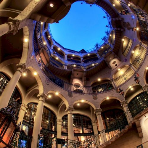 An ornate indoor courtyard with curved balconies and arches, lit by warm lights, framing a blue sky in a circular opening.