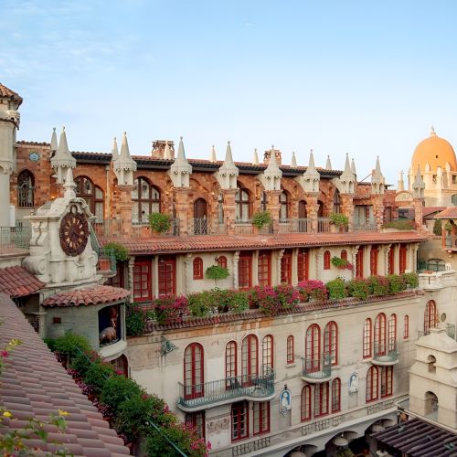 A grand Mediterranean-style hotel courtyard with arches, red doors/windows, domes, and blooming flowers under a blue sky.