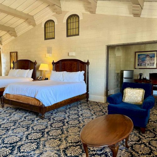 A spacious hotel room with two four-poster beds, a blue armchair, framed wall art, and a small coffee/serving area.