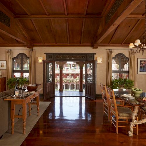 A warm dining room with wooden beams, a chandelier, and a set table beside an open archway to the courtyard.