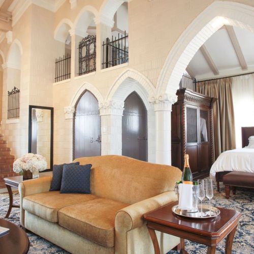 An elegant hotel suite interior with a chandelier, arched doorways, a plush orange sofa, and a lavish bed in the background.
