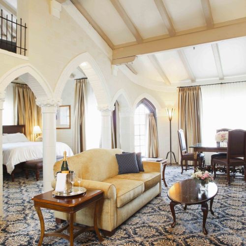 Elegant hotel suite with white arches, a yellow sofa, a coffee table with champagne, fireplace mantel, chairs, and sunlit windows.