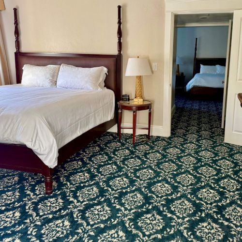 A hotel bedroom with a made bed, nightstand, lamp, patterned carpet, and an open door to a second bed.