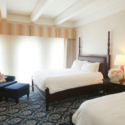 A bright hotel bedroom with two beds, white linens, striped curtains, bedside lamps, and upholstered seating.