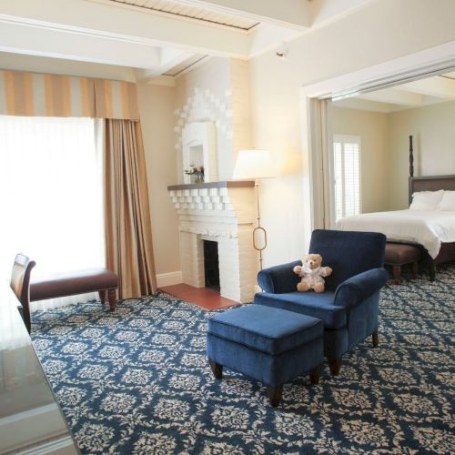 Bright, elegant bedroom with a four-poster bed, blue upholstered seating, patterned rug, curtains, and a white brick fireplace.