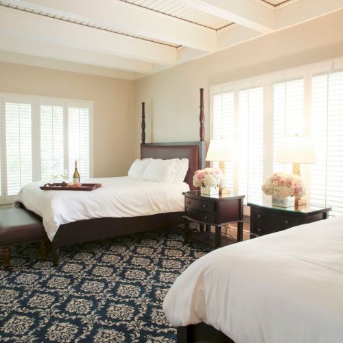 A bright, cozy bedroom with two beds, white linens, floral nightstands, lamps, and tall window blinds.