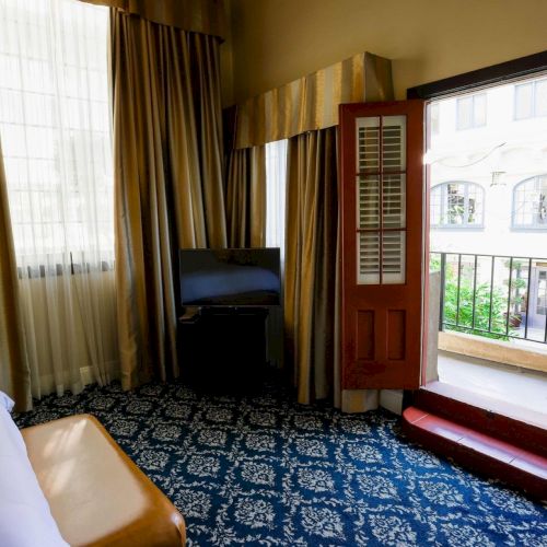 A hotel bedroom with a bed, TV stand, dark curtains, blue patterned carpet, and open doors/windows to a balcony.