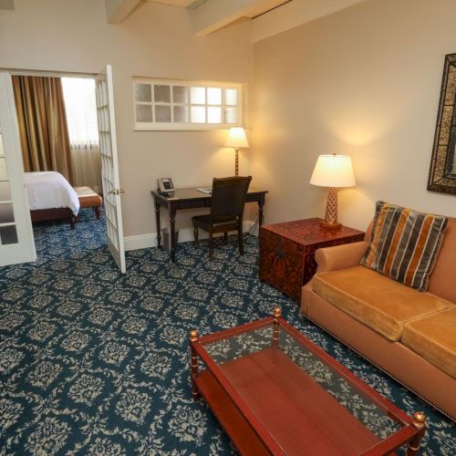 A hotel suite lounge with a tan sofa, blue patterned carpet, two lamps, desk, and framed mirror above a TV stand.
