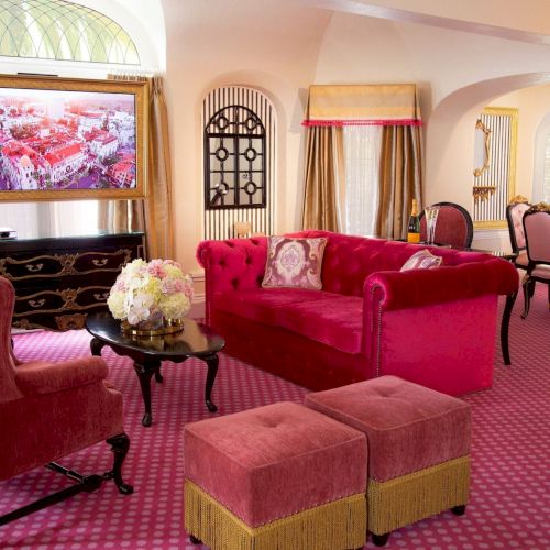 A luxurious lounge with a red velvet sofa, pink chairs and ottoman, ornate decor, framed artwork, curtains, and a dining area.