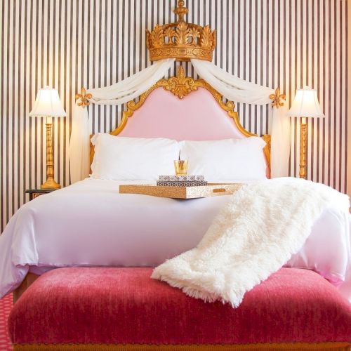 A luxurious bedroom with a gold crown headboard, pink drapes, striped wall, and a neatly made white bed with candles.