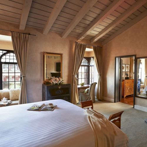 A bright, elegant bedroom with a large bed, arched windows and curtains, wooden beams, a TV, and a mirrored dresser.