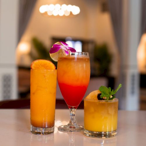 Three colorful cocktails in glasses&mdash;orange, red, and yellow&mdash;garnished with citrus and a flower on a bar counter.