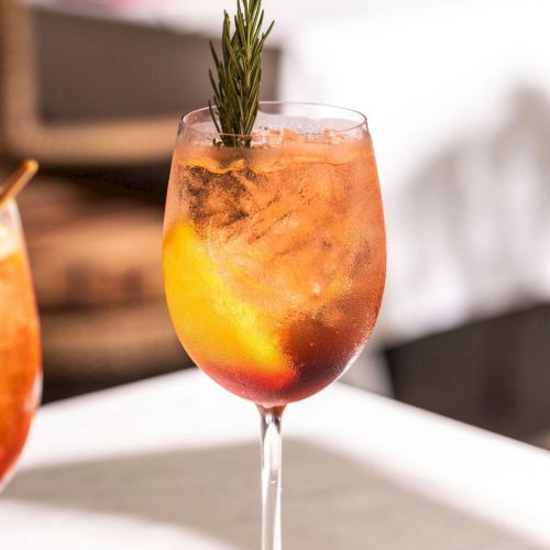 A stemmed glass holds an orange-red cocktail, topped with a rosemary sprig, on a table with soft white background bokeh.