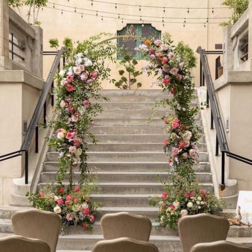 A floral wedding backdrop lines a stairway with hanging string lights and colorful roses, greenery, and chairs in front.