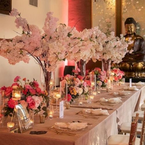 A decorated wedding banquet table with pink flower trees, candles, and a gold Buddha statue in the background.