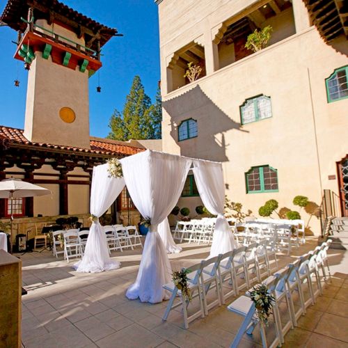 Outdoor wedding ceremony setup in a courtyard with white drapes, rows of chairs, and a sunny white building.