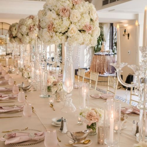 A lavish wedding reception table set with tall candles, crystal glassware, and abundant white-and-pink floral centerpieces.