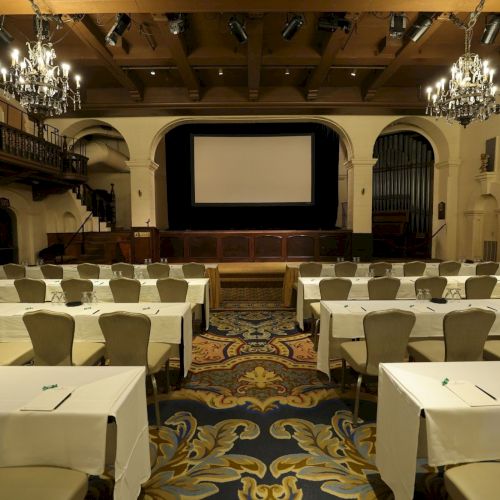 A grand conference hall with chandeliers, banquet tables, chairs, and a stage area with an empty projector screen.