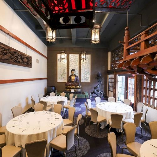 A formal restaurant dining room with white-table settings, a seated golden Buddha statue, and red-black decor overhead.