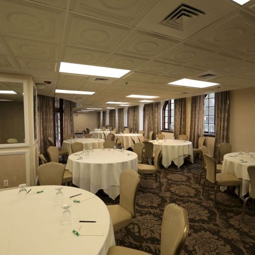An elegant conference room set up for a banquet, with round tables, beige chairs, white tablecloths, and large windows.