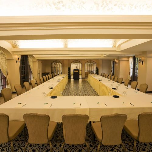 Elegant conference room with long banquet tables, chairs, and a patterned carpet leading to a central door, with equipment on the tables.
