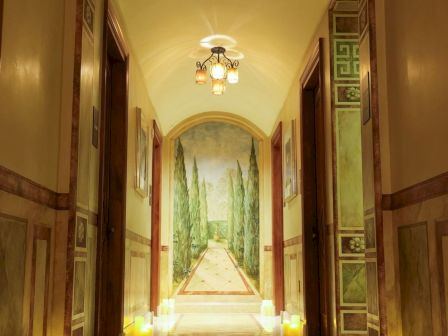 An ornate indoor hallway with wall art, a chandelier, warm ceiling lights, and many glowing candles lining the floor.