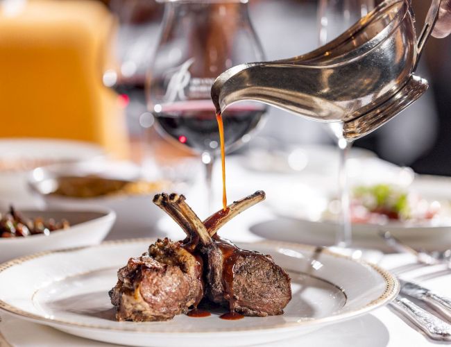 A chef pours rich sauce from a pan onto plated grilled lamb chops, finishing a restaurant-style dish with gravy drizzling down.