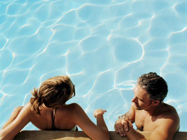 Two people lounge at a sunny pool, leaning on the edge as bright blue water ripples surround them.