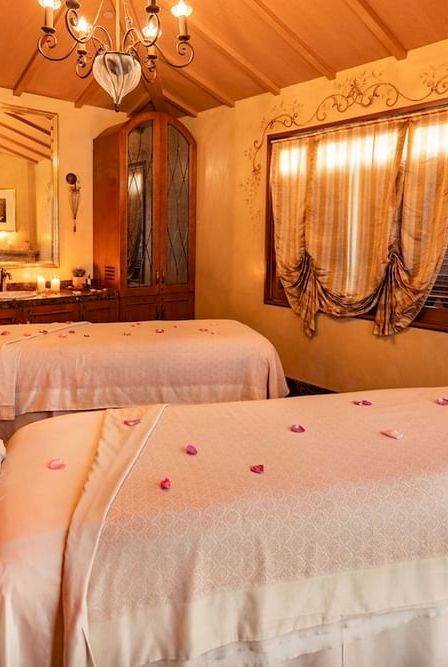 Cozy spa treatment room with two massage beds, rose petals, candles, and warm chandelier lighting.