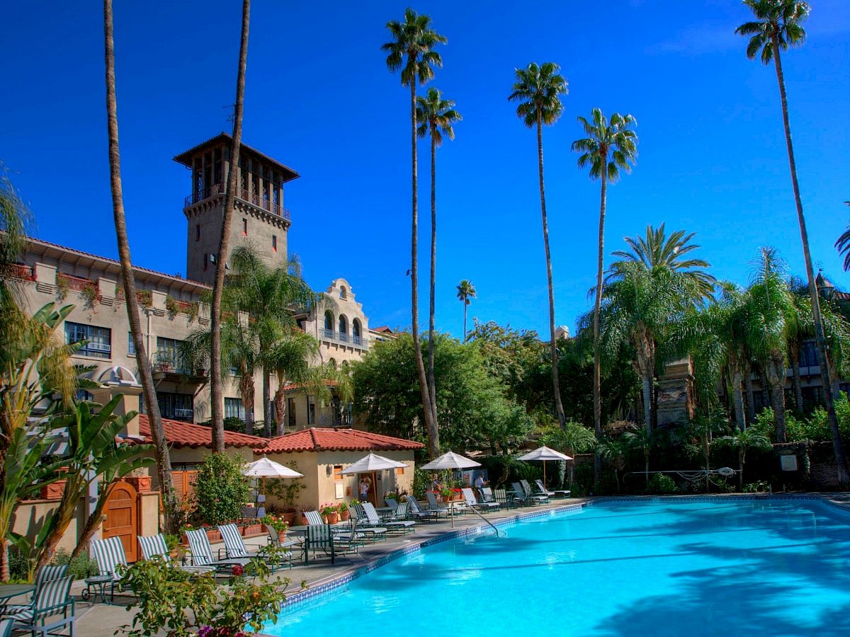 A sunny resort scene with a bright blue swimming pool, patio umbrellas, and tall palm trees against a clear blue sky.