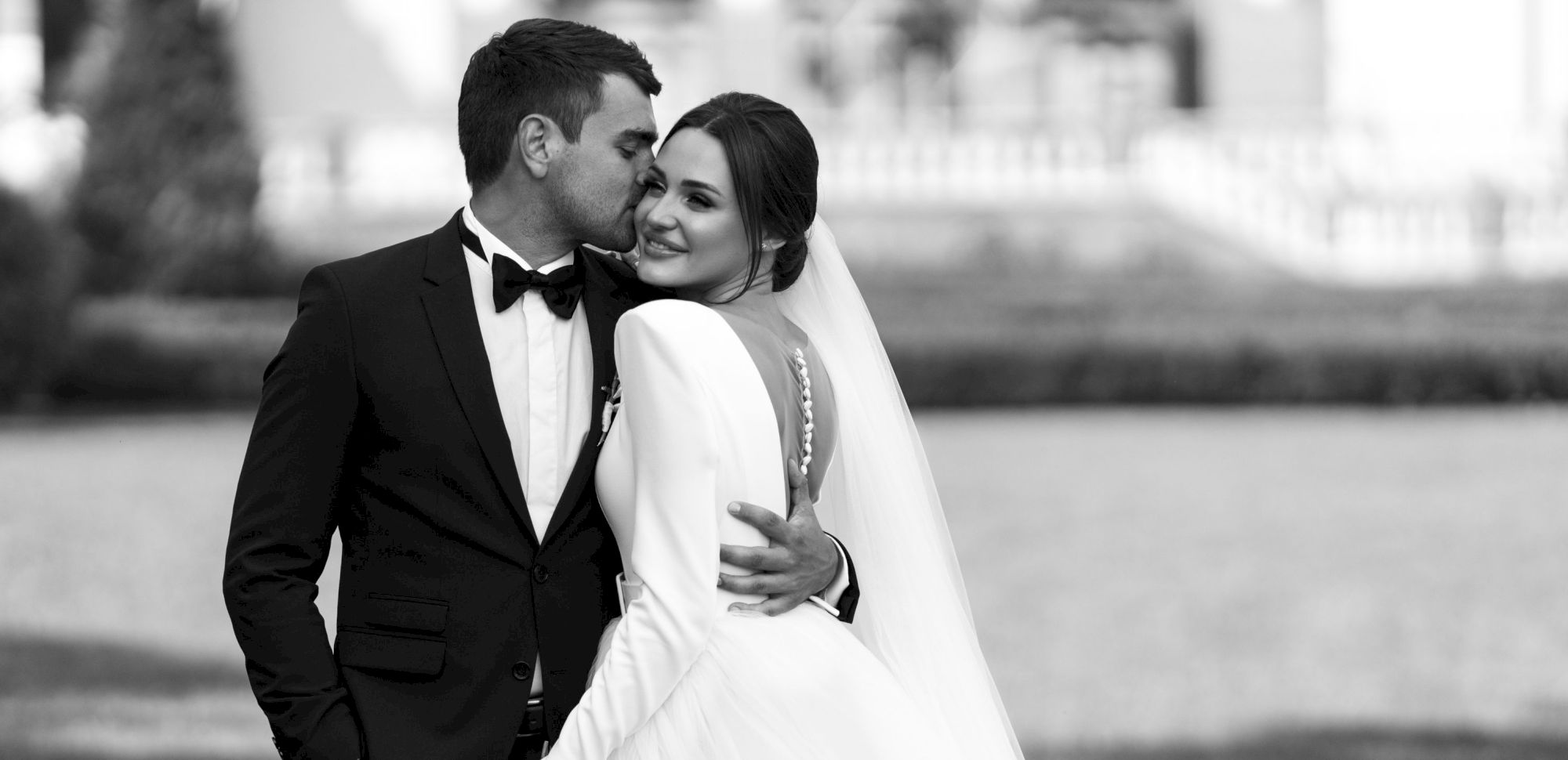 A bride and groom in black-and-white share a kiss outdoors, dressed in wedding attire.
