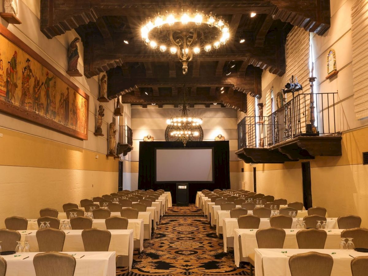 A banquet hall set for an event, with rows of tables, elegant chandeliers, framed artwork, and a projector screen at the front.