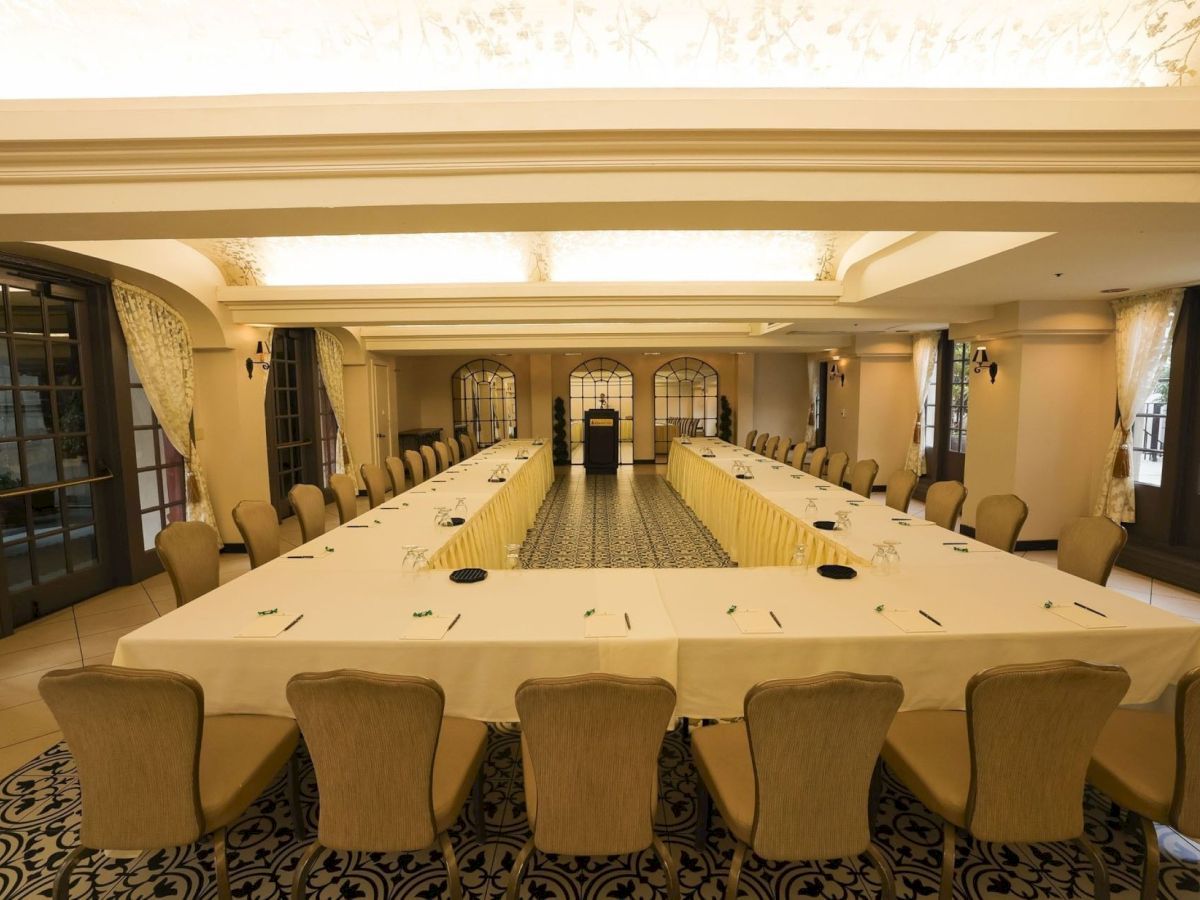 A formal banquet room with long tables, chairs, soft curtains, and warm lighting leading to a central podium at the far end.