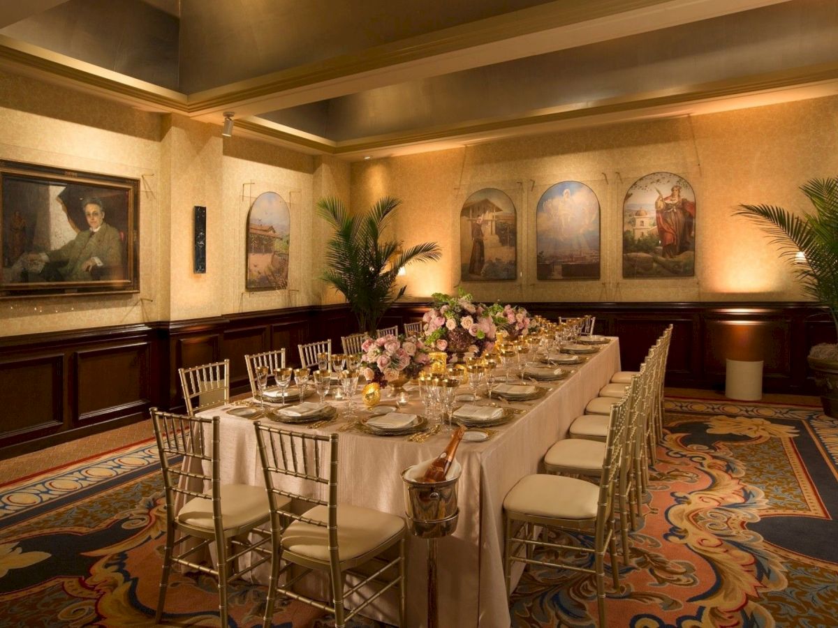 A formal banquet room with a long table set for dining, floral centerpieces, gold chairs, and wall art, under warm lighting.