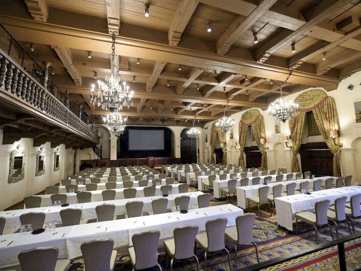 A grand conference hall with rows of banquet tables, chandeliers, and gold-and-green curtains.