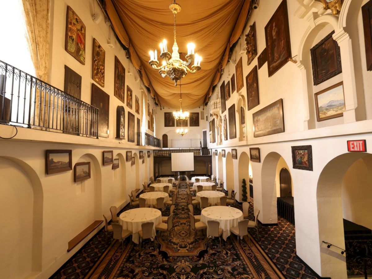 An elegant banquet hall with chandeliers, draped ceiling, framed paintings, arched corridors, and set dining tables for guests.