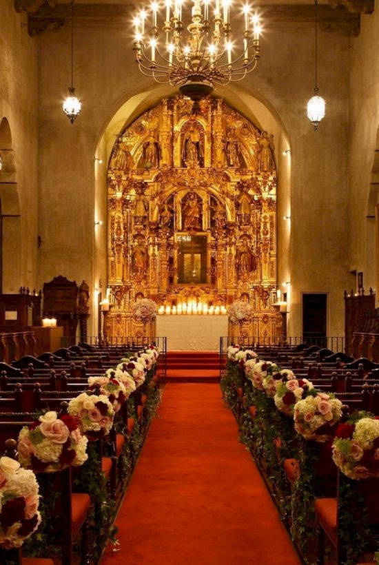A grand church interior with stained-glass windows, an ornate golden altar, chandeliers, and an aisle lined with flower arrangements.