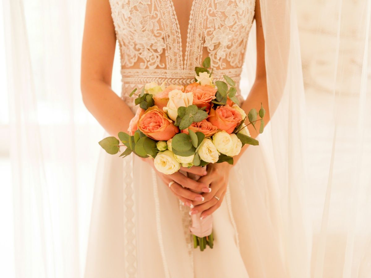 A bride in a white lace dress holds a bouquet of peach and cream roses with eucalyptus leaves.