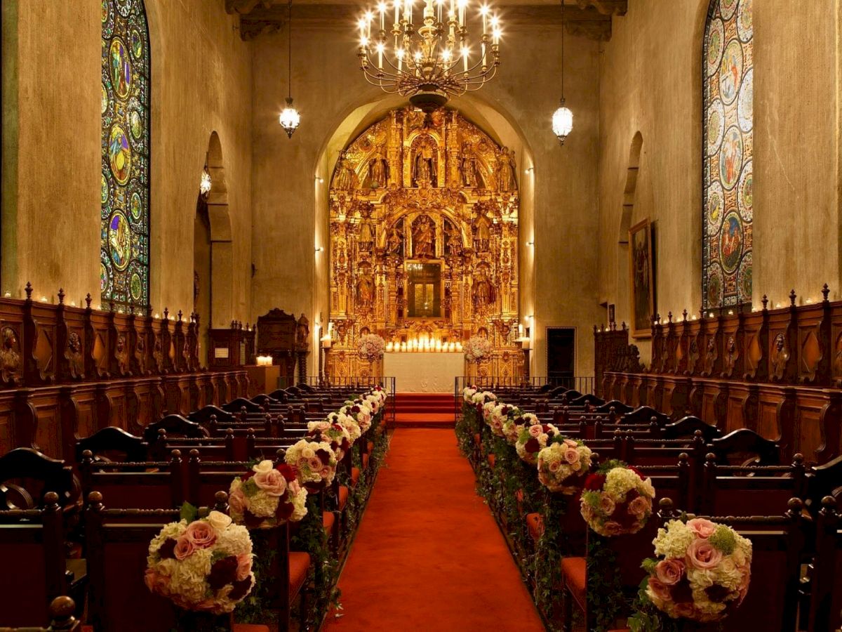 An ornate church interior with a golden altarpiece, sparkling chandelier, stained-glass windows, and floral arrangements along the aisle.