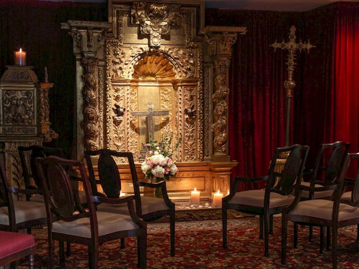 An ornate altar with gold carvings, candles, red velvet curtains, and chairs, with a bouquet at the center.