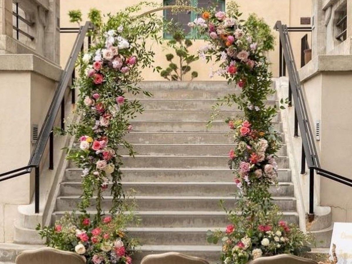 An indoor staircase is decorated with lush greenery, colorful flowers, and hanging string lights above, creating a wedding-like entryway.