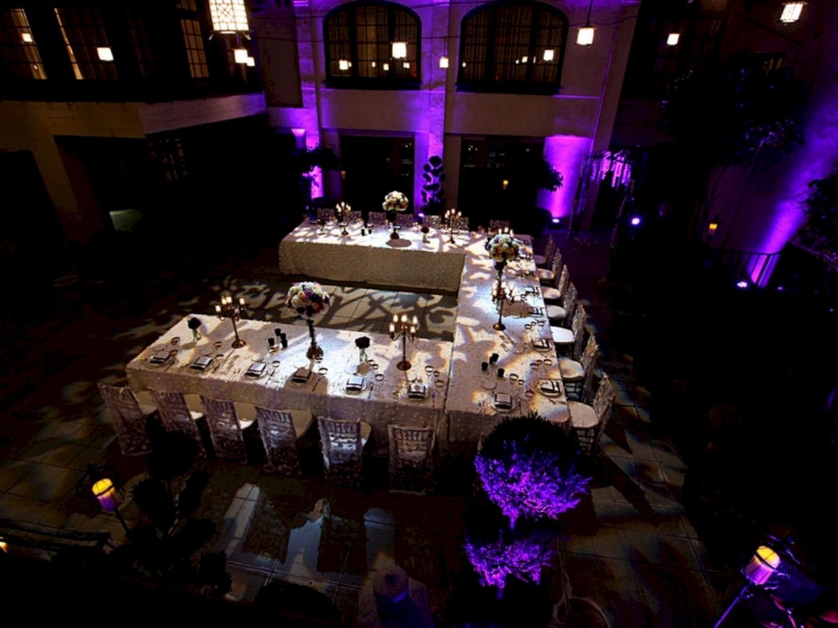 A banquet setup with a long table of candles and flowers in a courtyard, lit with purple stage lighting.