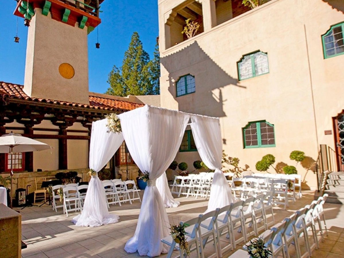 An outdoor wedding ceremony setup with white chairs, floral decor, and a draped arch in a sunny courtyard beside a stucco building.